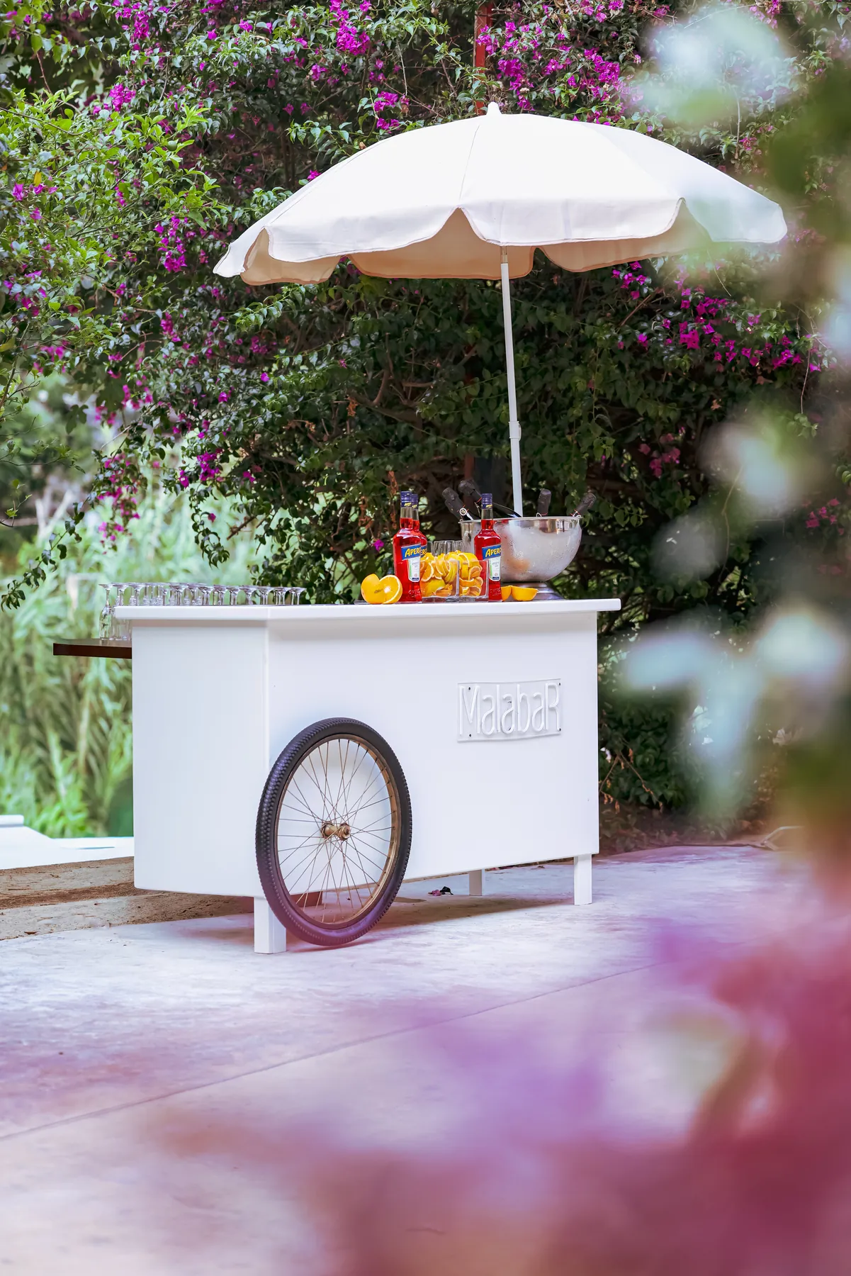 A white mobile drinks cart with a parasol in a flower-filled garden