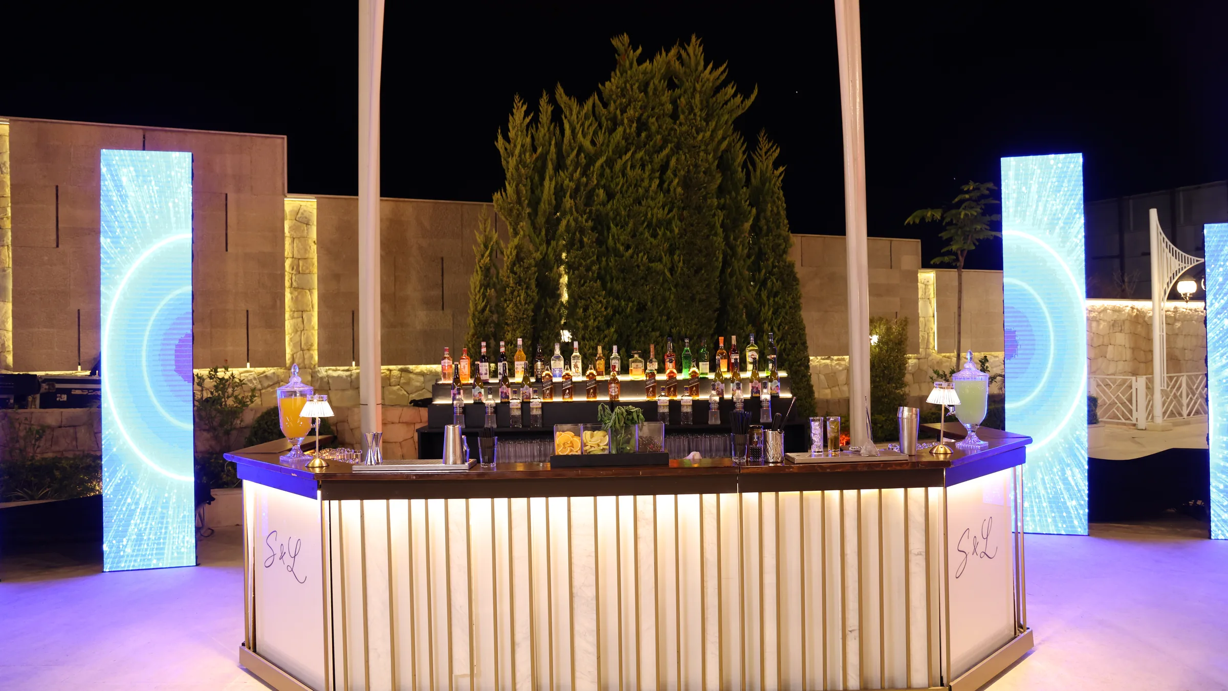 An illuminated Malabar bar with fluted panels, lit cypress trees and light columns at an evening celebration
